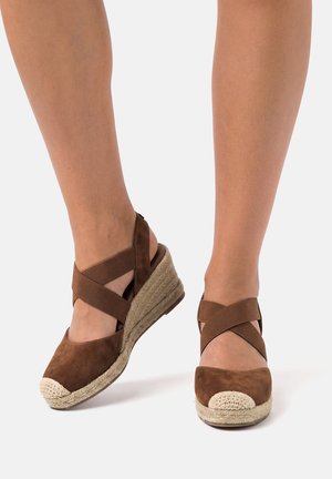 Alpargatas cuña marrones con correas elásticas cruzadas, usadas en pies desnudos sobre un fondo blanco liso.