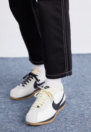 Osoba nosząca białe buty sportowe Nike z czarnym logo Swoosh, białe skarpetki oraz czarne spodnie z białym przeszyciem stojąca na niebieskim dywanie.