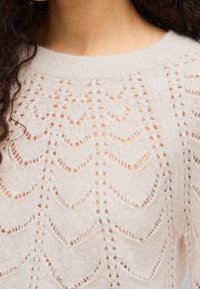 Κοντινή λήψη ενός λευκού πλεκτού πουλόβερ με σχέδια ζιγκ-ζαγκ και δαντέλα, φορεμένο από ένα άτομο με σκούρα σγουρά μαλλιά.