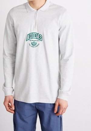 Grå langærmet polo med halvlukning og grønt broderet Adidas-logo på brystet. Stoffet ser blødt og struktureret ud.