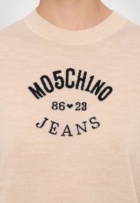 Camiseta de algodão bege clara com texto bordado em preto: "MO5CH1NO 86♥23 JEANS", gola redonda com detalhes em malha. Texto suave.
