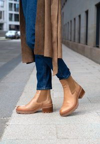 Stivaletti in pelle marrone chiaro con pannelli laterali elasticizzati, suole in gomma testurizzata e tacco impilato. Indossati con jeans in denim blu su un marciapiede.