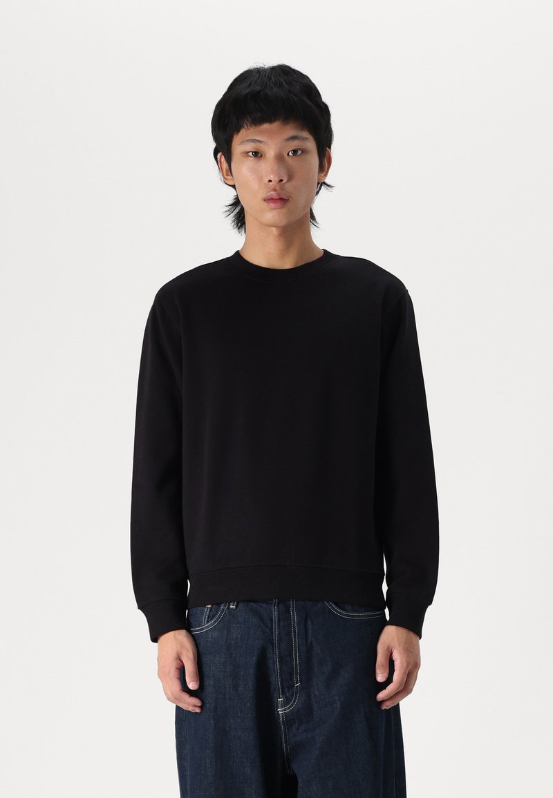 Sudadera negra de manga larga, con puños y dobladillo acanalados, diseño sencillo, fabricada en tejido suave, combinada con pantalones vaqueros oscuros de corte holgado.