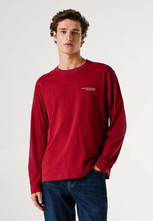 Rood T-shirt met lange mouwen van zachte stof en een ronde hals, met witte tekstborduursel op de linkerborst. Gecombineerd met een blauwe spijkerbroek.