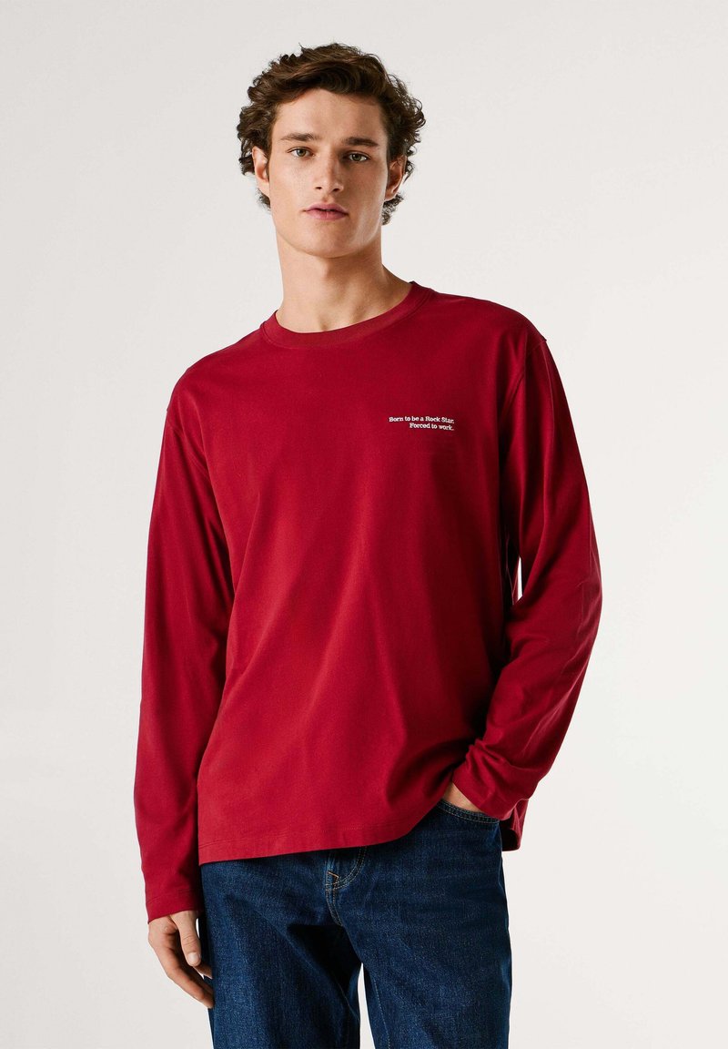 Maglietta rossa a maniche lunghe in tessuto morbido con scollo tondo, decorata con ricamo di testo bianco sul petto a sinistra. Abbinata a jeans blu.