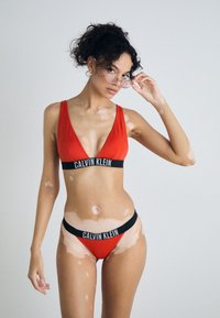 Crveni bikini set s dubokim izrezom i crnim brendiranim pojasom. Izrađen od glatkog materijala, naglašava minimalistički dizajn.