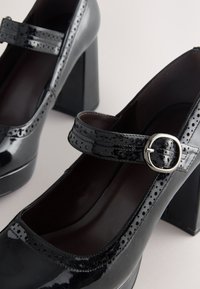 Black patent leather Mary Jane heels with brogue detailing, a rounded toe, a silver buckle strap, and thick block heels on a light background.