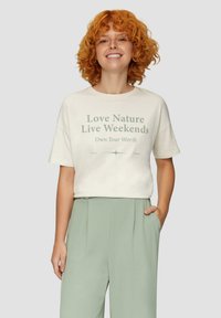Crème katoenen t-shirt met korte mouwen, met groene tekst: "Hou van de natuur Leef in het weekend Wees je eigen waarde." Model draagt lichtgroene broek.