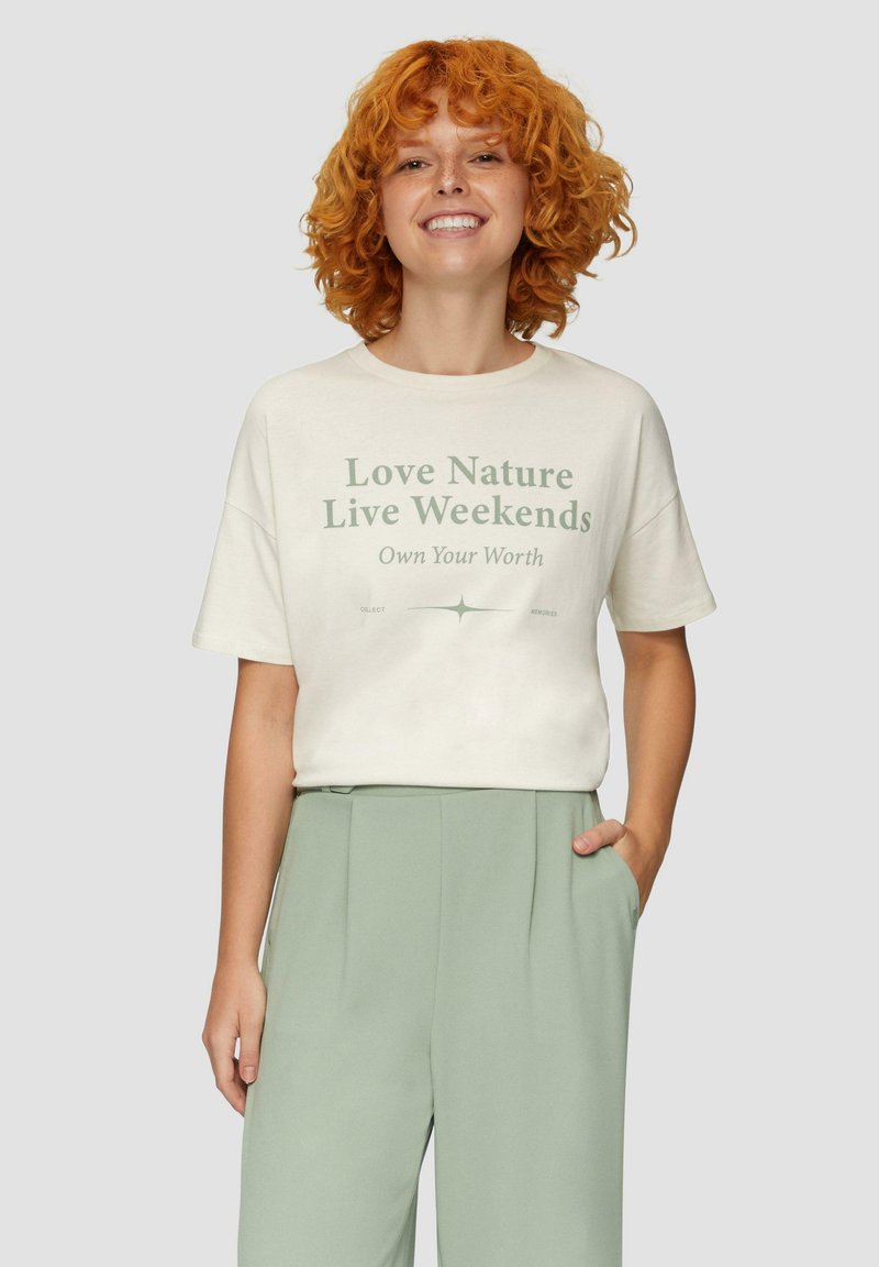 Crème katoenen t-shirt met korte mouwen, met groene tekst: "Hou van de natuur Leef in het weekend Wees je eigen waarde." Model draagt lichtgroene broek.