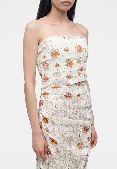 Femme portant une robe blanche bustier avec des motifs floraux multicolores et des pois dorés, debout devant un fond clair et uni.
