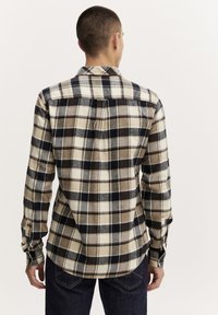 Het shirt heeft een ruitpatroon in beige, bruin en marineblauw, met een button-down kraag en lange mouwen, gemaakt van zachte stof.