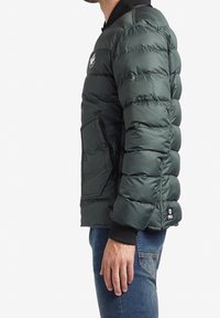 Zimní bunda v tmavě zelené barvě, se zipem vpředu, elastickými man�žetami a postranními kapsami; hladká textura s prošívaným vzorem.