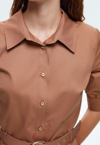 Chemise marron à boutons avec un col pointu, des manches courtes bouffantes et une ceinture beige avec une boucle circulaire. Tissu lisse et léger.