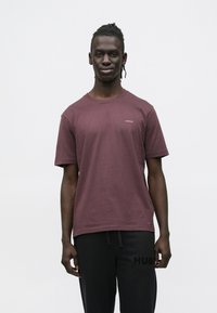 Burgundska bombažna majica z okroglim izrezom in kratkimi rokavi; na levi strani prsi je majhen logotip "HUGO". Model nosi črne trenirke.