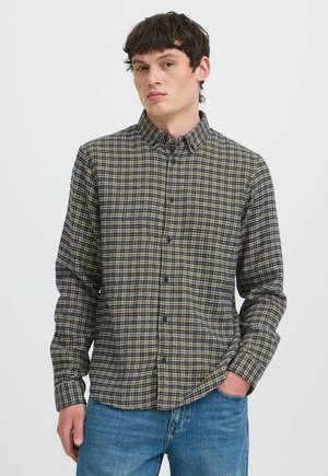 Jonge man met een dichtgeknoopte geruite lange mouwen blouse en blauwe spijkerbroek, staand met één hand in de zak tegen een effen achtergrond.
