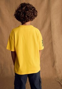 Geel kortegener t-shirt gezien vanuit de achterkant, met een ronde halslijn en een logo accent op de linker mouw. Casual pasvorm.