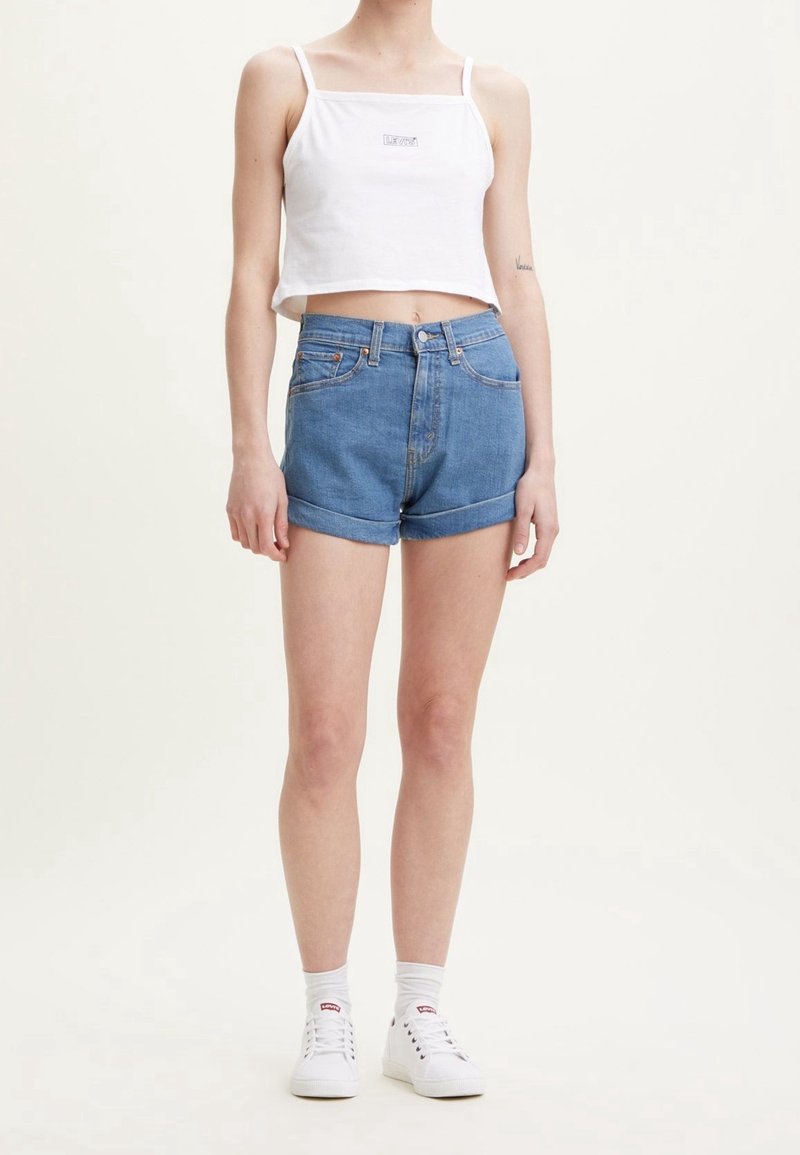 Personne portant un débardeur court blanc, un short en jean bleu à ourlet roulé, des chaussettes blanches et des baskets blanches, debout devant un fond uni.