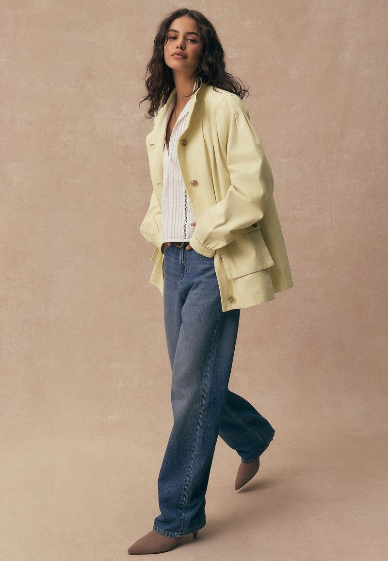 Femme aux longs cheveux bouclés portant une veste jaune pâle, un chemisier blanc, un jean large bleu et des chaussures pointues marron, debout les mains dans les poches.