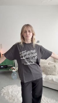 Νεαρή γυναίκα με ξανθά μαλλιά, ντυμένη με μαύρο μπλουζάκι με στάμπες και παντελόνι, στέκεται μέσα σε ένα ζεστό καθιστικό με τα χέρια ανοιχτά.