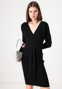 Vestido de malha preto com um decote em V profundo, mangas longas e um cinto laço. Acentuado com uma pequena bolsa de mão texturizada prateada.