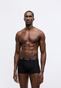 Fekete boxer short, Puma logóval ellátott derékrésszel, testhez simuló dizájnnal, sima textúrával, bemutatva egy izmos férfi modellen, semleges háttér előtt.
