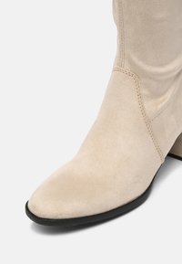 Botín de ante beige hasta el tobillo con punta afilada, superficie texturizada y un tacón bajo y apilado. Detalle de costura visible a lo largo de la costura lateral.