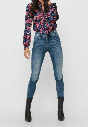 Femme portant un jean skinny bleu délavé taille haute, des bottines noires et un chemisier à manches longues à fleurs roses et bleues.