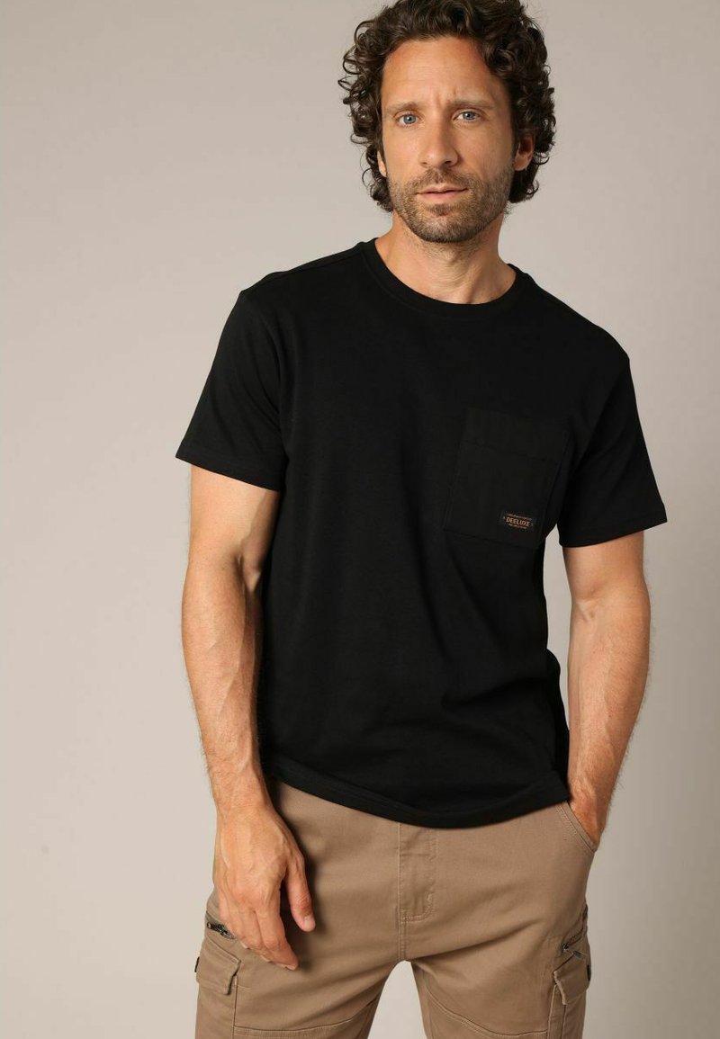 T-shirt en coton noir avec un col rond et des manches courtes. Il comporte une poche avant avec une étiquette, associé à un short cargo beige.