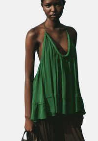 Haut halter vert en tissu léger, avec un décolleté en V profond, coupe ample et texture plissée, associé à un bas noir.