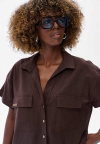 Kobieta o kręconych włosach, nosząca duże okulary w muszelkowym stylu, srebrne kolczyki w kształcie kółek oraz ciemnobrązową koszulę z guzikami i kieszeniami.