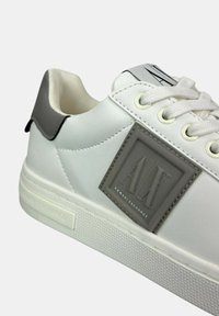 Sneaker bianco con accenti grigi, realizzato in materiale sintetico liscia. Presenta una suola texturizzata e un logo ben in evidenza sul lato.