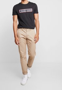 Černé tričko s krátkým rukávem s bílým logem "HILFIGER". Světle béžové chinos kalhoty s ohrnutými manžetami. Bílé sportovní boty s jemnými značkovými detaily.