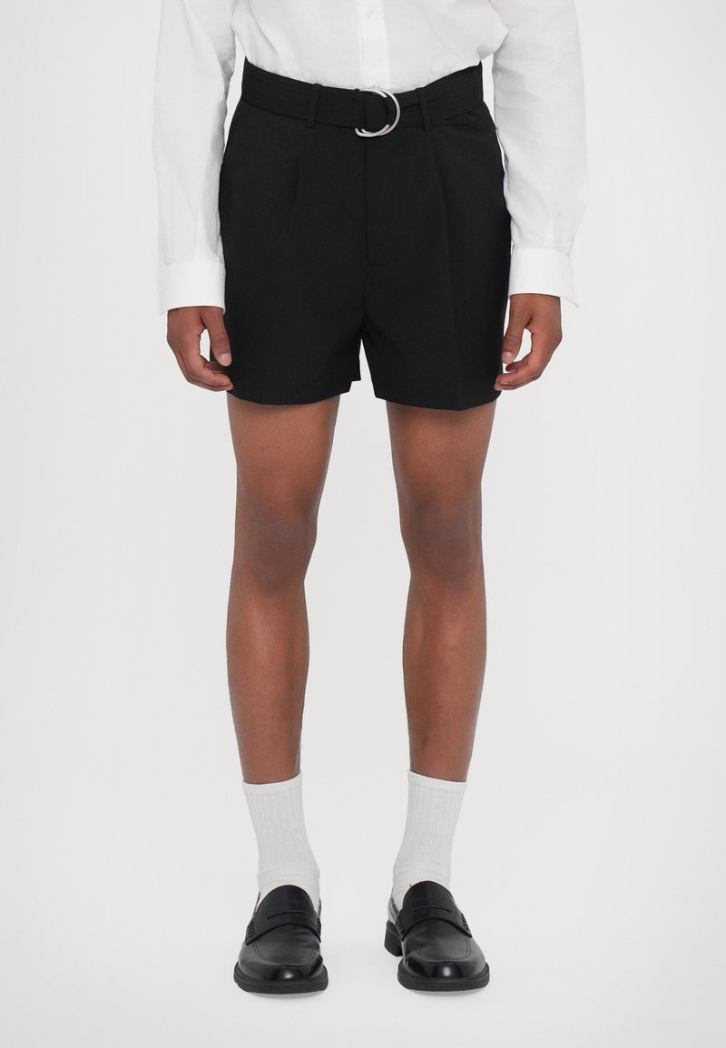 Fekete, fazonra szabott sort övvel, fémből készült, kör alakú csattal, fehér zoknikkal és fekete slip-on cipőkkel párosítva.