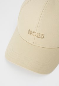 Lys beige baseball cap laget av glatt stoff, med en sydd logo "BOSS" i matchende tråd, synlige sømmer og en buet skjerm.
