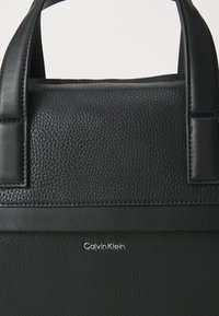 Black leather handbag with a textured surface and smooth accents. Features a top handle and embossed "Calvin Klein" logo.