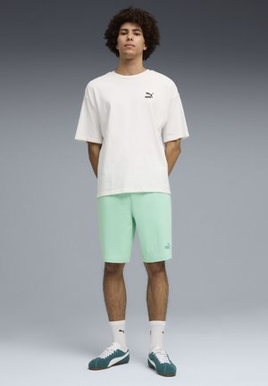 Joven con cabello rizado que lleva una camiseta blanca de Puma, pantalones cortos verde menta, calcetines blancos de Puma y zapatillas turquesas, de pie frente a un fondo gris.