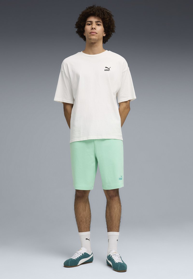 Jeune homme aux cheveux bouclés portant un t-shirt blanc Puma, un short vert menthe, des chaussettes blanches Puma et des baskets bleu sarcelle, debout devant un fond gris.