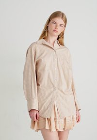 Jonge vrouw met lang blond haar, gekleed in een oversized beige geruit button-up shirt en een lichte gelaagde rok, staand tegen een effen achtergrond.
