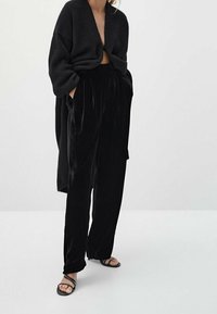 Black, oversized knit cardigan with dropped shoulders, paired with high-waisted black velvet trousers featuring pleats. Accentuated with black sandals.