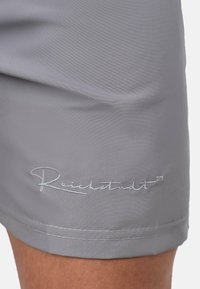Shorts gris en tissu lisse avec une texture subtile, comportant un logo brodé "Reichstadt" en blanc sur l'ourlet inférieur.
