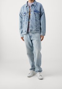 Giacca di jeans azzurro chiaro con due tasche frontali, abbinata a jeans azzurro chiaro. Sneakers bianche completano l'outfit con una t-shirt grafica viola sotto.