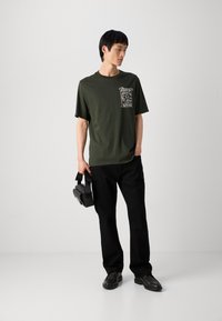Mørkegrøn bomulds t-shirt med et grafisk print på venstre bryst, parret med sorte bukser og sorte sko. Modellen holder en taske.