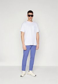 Vit bomulls-t-shirt med en liten ficka, ljusblå byxor och vita sneakers. Modellen bär svarta solglasögon och står mot en neutral bakgrund.