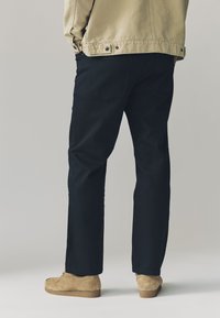 Sorte bukser lavet af slidstærkt stof, med lige ben, kombineret med tanfarvede ruskindssko, og med en enkel lomme detalje.