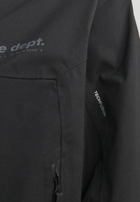 Schwarze Jacke aus weichem Stoff, mit einer Reißverschlusstasche, schrägen Nähten und einem Druckdesign, das "dept." mit dem Detail "TECH FUSION" zeigt.