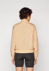 Veste bomber légère en tissu beige doux, avec un ourlet et des poignets côtelés. Le dos présente un col montant et une texture lisse.