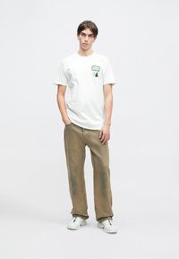 Bílé bavlněné tričko s grafickým motivem na hrudi, v kombinaci s volnými béžovými kalhotami a bílými teniskami.