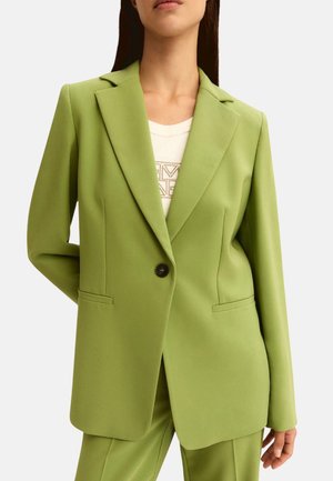 Donna che indossa un blazer verde chiaro su misura con un solo bottone nero sopra una maglietta bianca con un motivo decorativo.