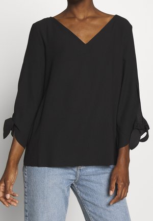 Schwarze V-Ausschnitt-Bluse mit 3/4-Ärmeln, die mit Schleifenverzierungen an den Ärmelöffnungen versehen ist. Glatte Textur und lockere Passform. Kombiniert mit hellblauen Jeans.