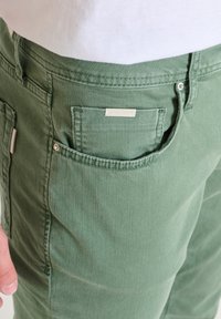 Pantalones de algodón verdes con un bolsillo que presenta una etiqueta de logo plateada. La tela tiene una textura suave y un sutil detalle de costura a lo largo de las costuras.